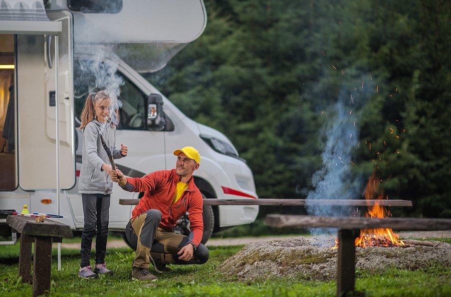 A father and daughter enjoy cooking over an open fire in front of their RV campsite. 